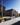 Sunny exterior visualization of a modern, multi-story brick apartment building on a city corner, featuring a cobblestone sidewalk with flower planters, passing cars, and pedestrians.